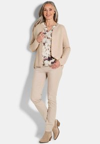 Cardigan en tricot beige sur une blouse à fleurs, associé à un pantalon skinny beige clair et des bottines tan. Textures douces, design simple, couleurs neutres.