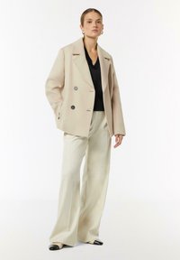 Beige Mantel mit doppelter Knopfreihe und großen schwarzen Knöpfen, getragen über einem schwarzen Oberteil und cremefarbenen, weit geschnittenen Hosen. Glatte Textur, maßgeschneiderter Schnitt.