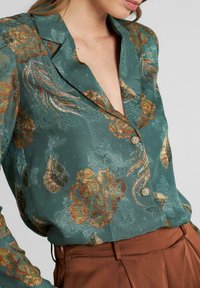 Blusa verde con patrones florales en tonos dorados y óxido, con cuello y cierre de botones. La tela es transparente y texturizada.