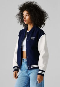 Kvinne med krøllete hår iført en Marineblå og hvit Red Bull Racing-jakke, hvit crop top og lyseblå jeans, stående sideveis.