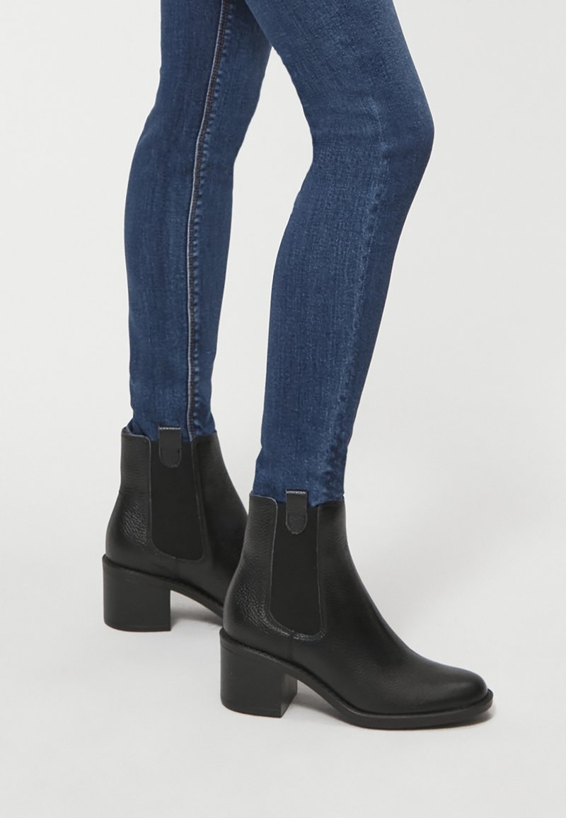 Bottines en cuir noir avec panneaux élastiques latéraux et talon carré, portées avec un jean skinny bleu foncé. Texture lisse avec des détails subtils.