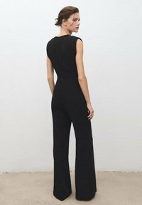 Monos de mujer negros con mangas tipo cap sleeve, corte ajustado, pierna ancha y textura suave. Presenta una cremallera oculta en la parte trasera y un diseño minimalista.