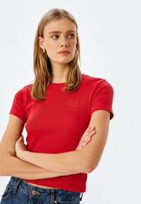 Rød ribbet t-shirt med korte ærmer, en tætsiddende design og et lille broderet logo på brystet. Båret med mørkeblå jeans.