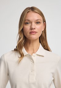 Beige polo shirt gemaakt van gestructureerde stof, met een kraag, vier knopen en een getailleerd ontwerp, gedragen door een persoon met lang haar.