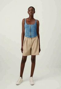 Woman wearing sleeveless blue denim zip-up top, beige pleated shorts, and white sneakers, standing against a plain background.