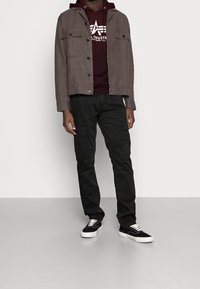 Uomo che indossa una giacca marrone sopra un felpa bordeaux con logo, pantaloni cargo neri e sneakers nere con suola bianca, in piedi contro un muro semplice.