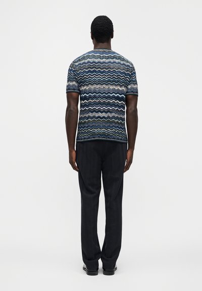 Cămașă cu mâneci scurte, cu model zigzag în nuanțe de albastru închis și multicolor, asortată cu pantaloni texturați închis la culoare și pantofi negri, privită din spate.