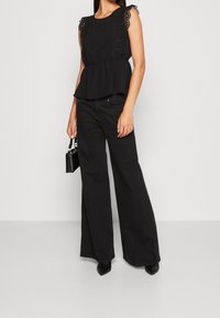 Haut noir à basque avec détails en dentelle sur les manches, associé à un jean noir à jambes larges. Modèle tenant un petit sac à main noir, portant des talons pointus.