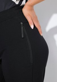 Svarta sweatpants med en slät textur, elastisk midja och en sidoficka med dragkedja för bekvämlighet. Närbilden framhäver detaljer.