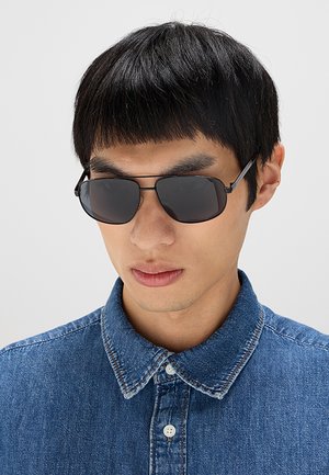 Giovane uomo con capelli neri lisci che indossa occhiali da sole neri e una camicia di jeans blu abbottonata su uno sfondo chiaro e uniforme.