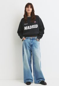 Čierna mikina s nápisom "MADRID" v bielej farbe a červenými akcentmi, skombinovaná s nohavicami s voľným strihom v svetlom modrom odtieni s vyblednutým vzhľadom.