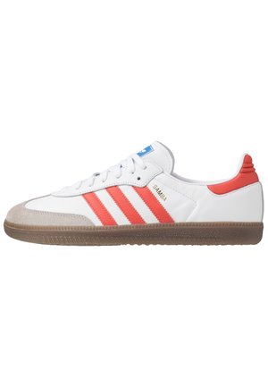Biały but sportowy Adidas Samba z czerwonymi paskami, brązową gumową podeszwą, zamszowym noskiem i niebieskim logo na języku, napis "SAMBA" po boku w kolorze złotym.