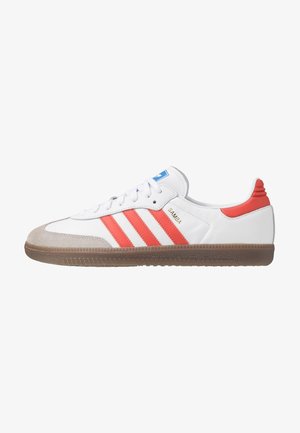 Biały but sportowy Adidas Samba z czerwonymi paskami, brązową gumową podeszwą, zamszowym noskiem i niebieskim logo na języku, napis "SAMBA" po boku w kolorze złotym.