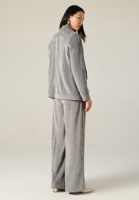 Completo in velluto a coste grigio con giacca lunga e pantaloni a gamba larga. Presenta una superficie testurizzata, colletto a rever e sottili strisce verticali.