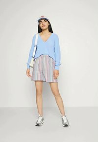 Jovem mulher de pé, vestindo um suéter azul em V, calções às riscas, sapatilhas prateadas, uma bolsa cruzada branca e um boné azul claro e rosa.