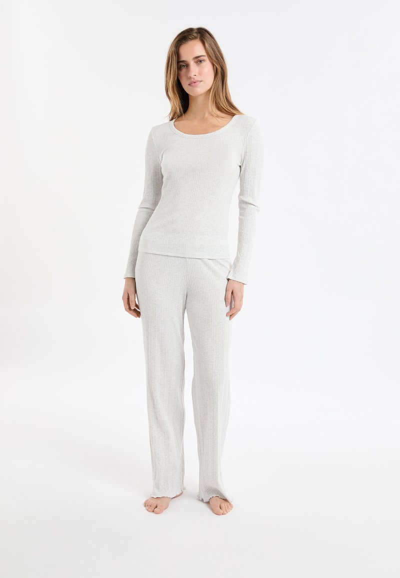 Kobieta stoi boso w jasnoszarej, prążkowanej bluzie z długim rękawem oraz dopasowanych szerokich spodniach na białym tle.