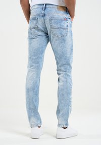 Šviesiai mėlynos džinsinės kelnės su tiesiomis klešnėmis, užpakaliniai kišenės su siuvinėjimo detalėmis ir ruda juostos etiketė. Dėvimos su baltomis sportinėmis bateliais.