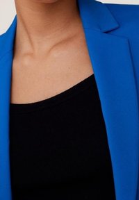 Modrý blejzr s vyříznutými klopami, v kombinaci s černým tílkem bez rukávů. Blejzr má hladkou texturu a střih na míru.