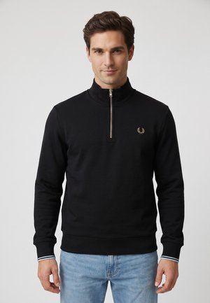 Hombre vistiendo un jersey negro con cremallera hasta el pecho, con un pequeño logo de laureles dorado y vaqueros azul claro, de pie frente a un fondo gris liso.