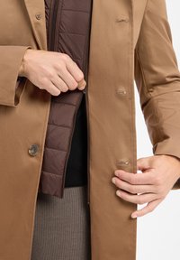 Hände, die eine braune, gestepfte Jacke unter einem hellen Überrock zippen und die Schicht- und Verschlussdetails der Oberbekleidung zeigen.