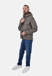 Giacca zip-up grigio scuro con cappuccio, dotata di due tasche sul petto e dettagli sulle spalle. Abbinata a jeans blu e sneaker bianche.