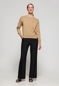 Beige Strickpullover mit Rundhalsausschnitt und langen Ärmeln, kombiniert mit hochgeschnittenen schwarzen Hosen. Sanfte Textur und lässige Passform.