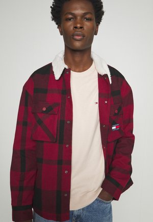 Camicia a quadri rossa e nera con colletto in pile bianco, dotata di due tasche frontali, chiusa con bottoni a pressione in metallo, indossata sopra una t-shirt a girocollo beige.