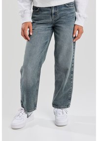 Jeans in denim blu chiaro, morbidi e dall'aspetto informale, con cuciture visibili, cinque tasche e una texture consumata, abbinati a scarpe da ginnastica bianche.