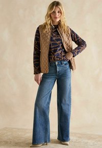 Beige gesteppte Weste über einem navyblauen gemusterten Oberteil, kombiniert mit weit geschnittenen blauen Jeans und beigen Absätzen vor einem strukturierten hellen Hintergrund.