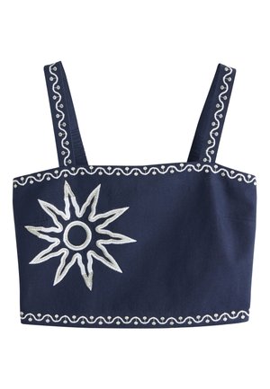 Femme aux longs cheveux blonds portant une tenue deux pièces bleu marine avec de grands motifs de soleil blancs, debout sur un sol rocheux contre un mur uni.