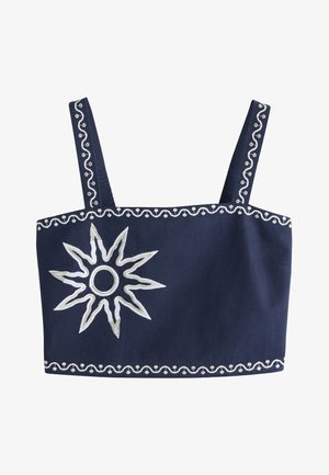 Femme aux longs cheveux blonds portant une tenue deux pièces bleu marine avec de grands motifs de soleil blancs, debout sur un sol rocheux contre un mur uni.