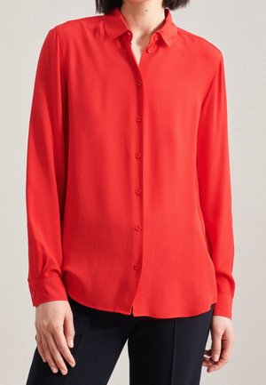 Kvinde iført en langærmet rød skjorte med knapper og krave, kombineret med sorte bukser, vist fra hals til hofter.