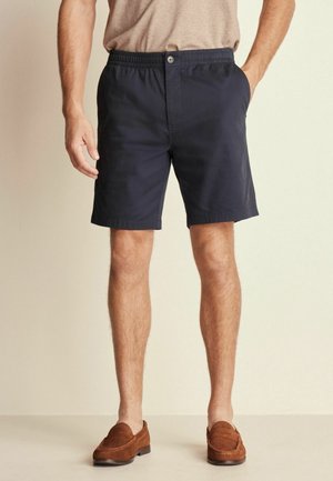 Homme portant un short bleu marine ajusté avec bouton et taille élastique, associé à des mocassins marron et une chemise beige.