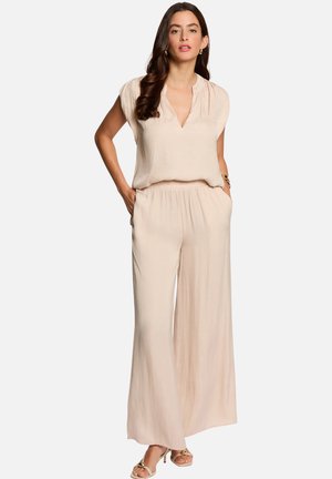 Femme aux longs cheveux foncés portant un chemisier beige à manches courtes et un pantalon large assorti, debout les mains dans les poches, fond neutre.