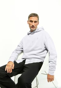 Haut à capuche violet clair, mélange de coton, poche kangourou, coupe décontractée. Porté par-dessus une chemise à carreaux blanche et grise. Pantalons noirs, assis sur une chaise.