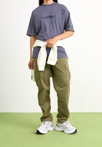 Camiseta gris con estampado gráfico, pantalones cargo oliva, sudadera blanca atada a la cintura y zapatillas deportivas blancas con acentos negros.