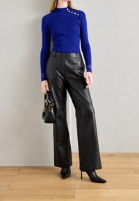 Femme portant un haut bleu à manches longues avec des boutons, un pantalon large en cuir noir, des bottes à talons hautes noires, tenant un sac à main noir.