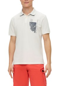 Weißes Baumwollpoloshirt mit kurzen Ärmeln, das auf der linken Seite ein blaues grafisches Design und einen kontrastierenden Kragen aufweist. Kombiniert mit roten Cargo-Shorts.