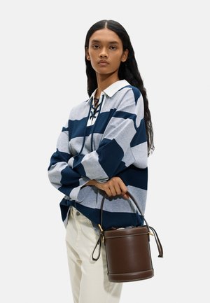 Kobieta z długimi ciemnymi włosami ubrana w niebiesko-szarą, paskowaną koszulę rugby, kremowe spodnie, trzymająca ciemnobrązową skórzaną torbę typu bucket.