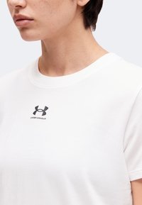 Persoon draagt een wit Under Armour T-shirt met kort donker haar, getoond van nek tot bovenste borst tegen een effen achtergrond.