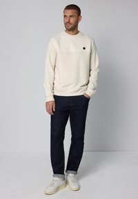 Světlobéžová mikina s texturovaným horním panelem a černým logem jako akcentem, doplněná tmavě modrými džínami a bílými teniskami.