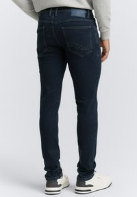 Dunkelblaue Jeans im Slim-Fit-Stil mit Gesäßtaschen und gewölbten Nähdetails, getragen mit weißen Sportschuhen.