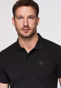 Junger Mann mit kurzen braunen Haaren und leichtem Bart, der ein schwarzes Poloshirt mit kleinem besticktem Logo auf der Brust trägt.