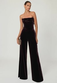 Ujjatlan overall gazdag bordó bársonyból, illeszkedő felsőrésszel, redőzött részletekkel és széles palazzo nadrággal. Kicsi textúrázott táska kiegészítő.