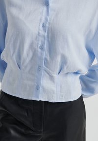 Chemise bleu clair boutonnée, rentrée dans un pantalon noir taille haute, mains posées dans le dos sur un fond uni.
