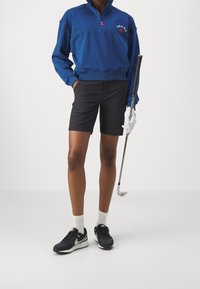 Sudadera azul con cremallera y logo rojo, pantalones cortos negros, zapatillas de deporte negras y blancas, y un golfista sosteniendo un palo. Calcetines blancos visibles.