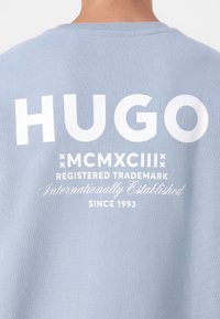 Világoskék pamut póló, nagy fehér felirattal, "HUGO," kiegészítő védjegy részletekkel és egy dekoratív 1993-as évszámmal alatta.