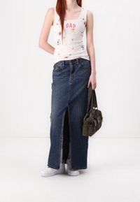 Débardeur floral blanc avec le texte "GAP", jupe maxi en denim bleu avec fente à l'avant, baskets blanches, portant un sac en velours côtelé vert olive.