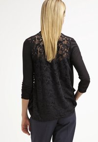 Parte superior de manga larga negra con un panel trasero de encaje que presenta patrones florales; tejido suave y corte holgado con dobladillo en forma de cucharada en la parte posterior.