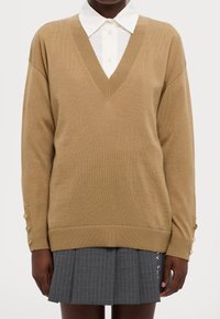 Pull beige à col en V en tissu tricoté, avec des poignets côtelés ornés de boutons et une coupe décontractée, porté sur une chemise blanche à col et une jupe plissée grise.